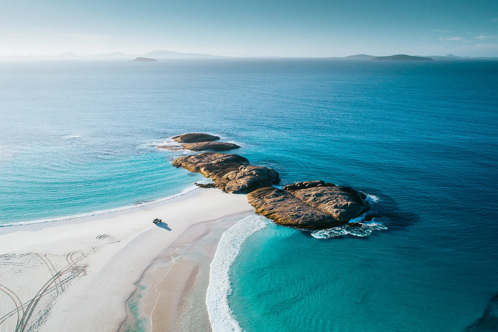 Aerial of granite outcrops on a white beach with a vehicle parked on the sand
