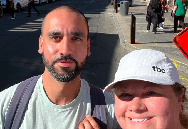 Two people pose for a selfie on a sunny street, one wearing a white cap and the other with a backpack