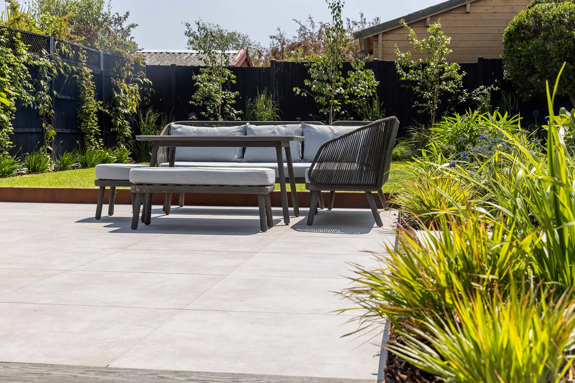 A stylish outdoor bench surrounded by greenery and plants on a paved surface in a sunny garden setting.