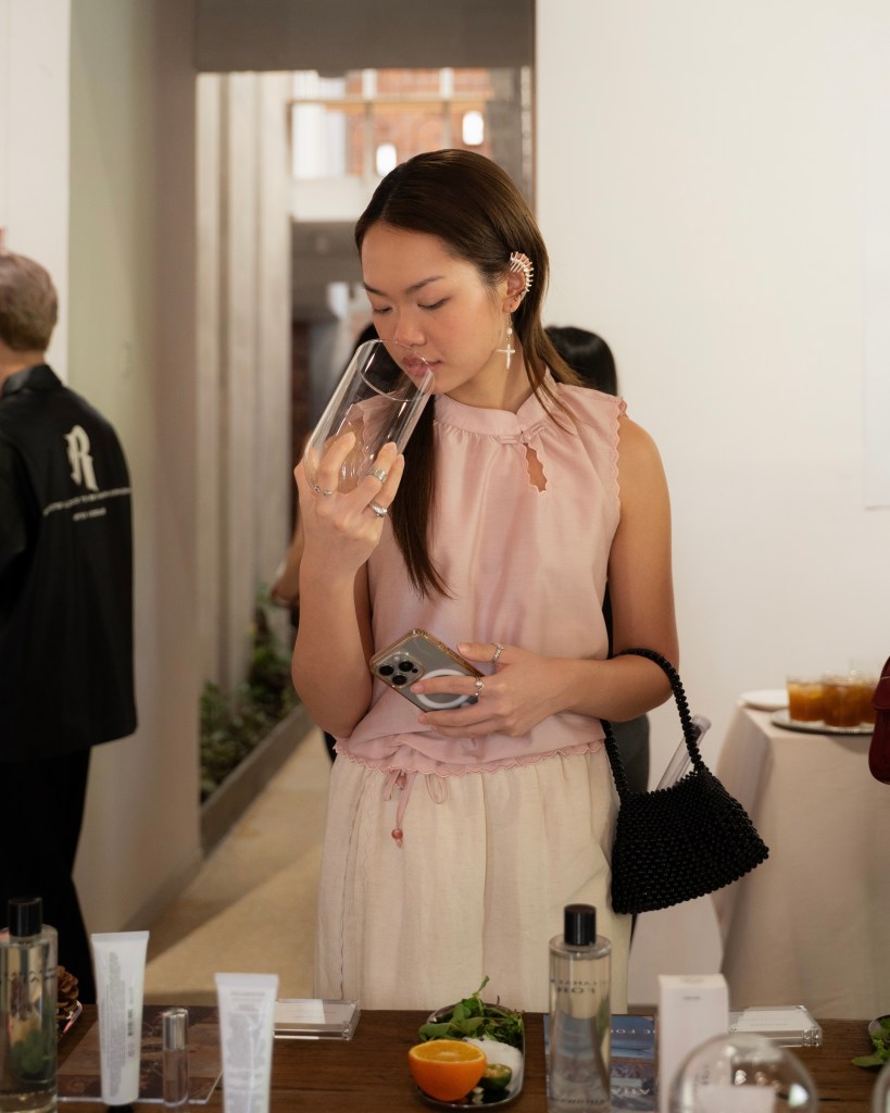 A young woman in a beige tank top holds a clear glass with her head tilted slightly, appearing to inspect its contents. She is in a stylish, modern setting with tables displaying beauty products and fresh ingredients.