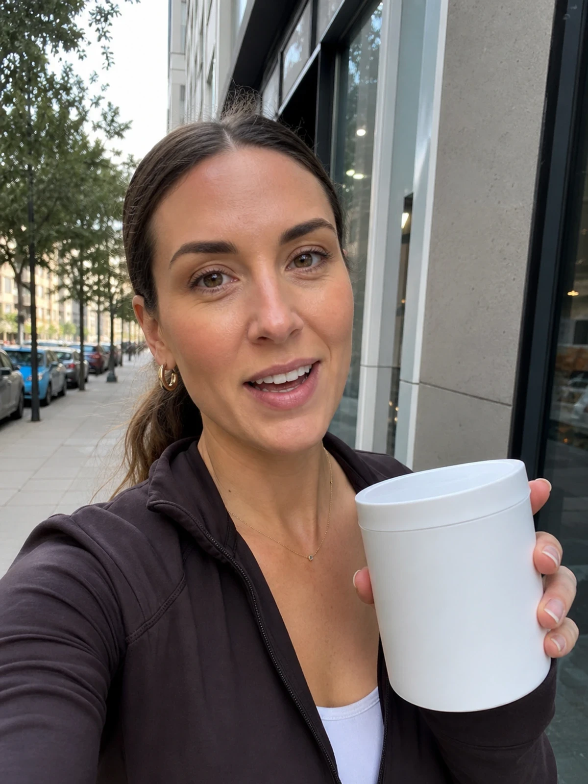 UGC-style photo: female creator in her 30s, on a city sidewalk, holding a plain wellness product while talking to camera, casual handheld angle.