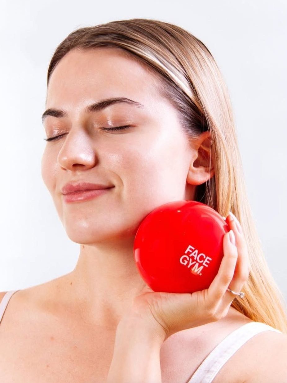 A woman with long hair is smiling while holding a red facial exercise ball labeled 'FACE GYM' against her cheek, suggesting a skincare routine focused on facial fitness.