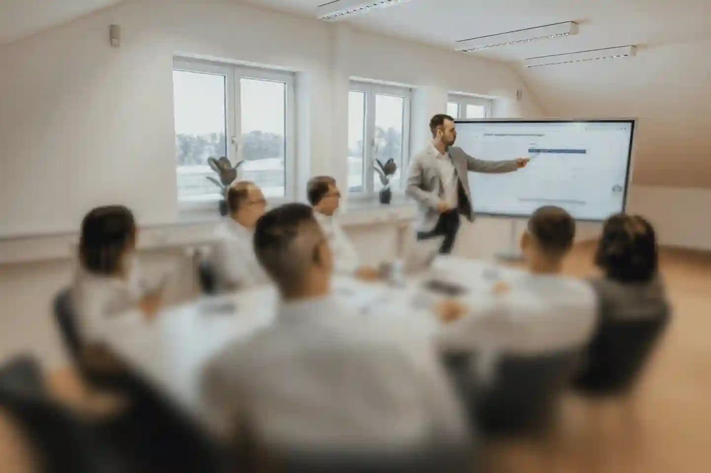 A business professional stands in front of a large screen giving a presentation to a group of colleagues seated around a conference table in a bright office setting.