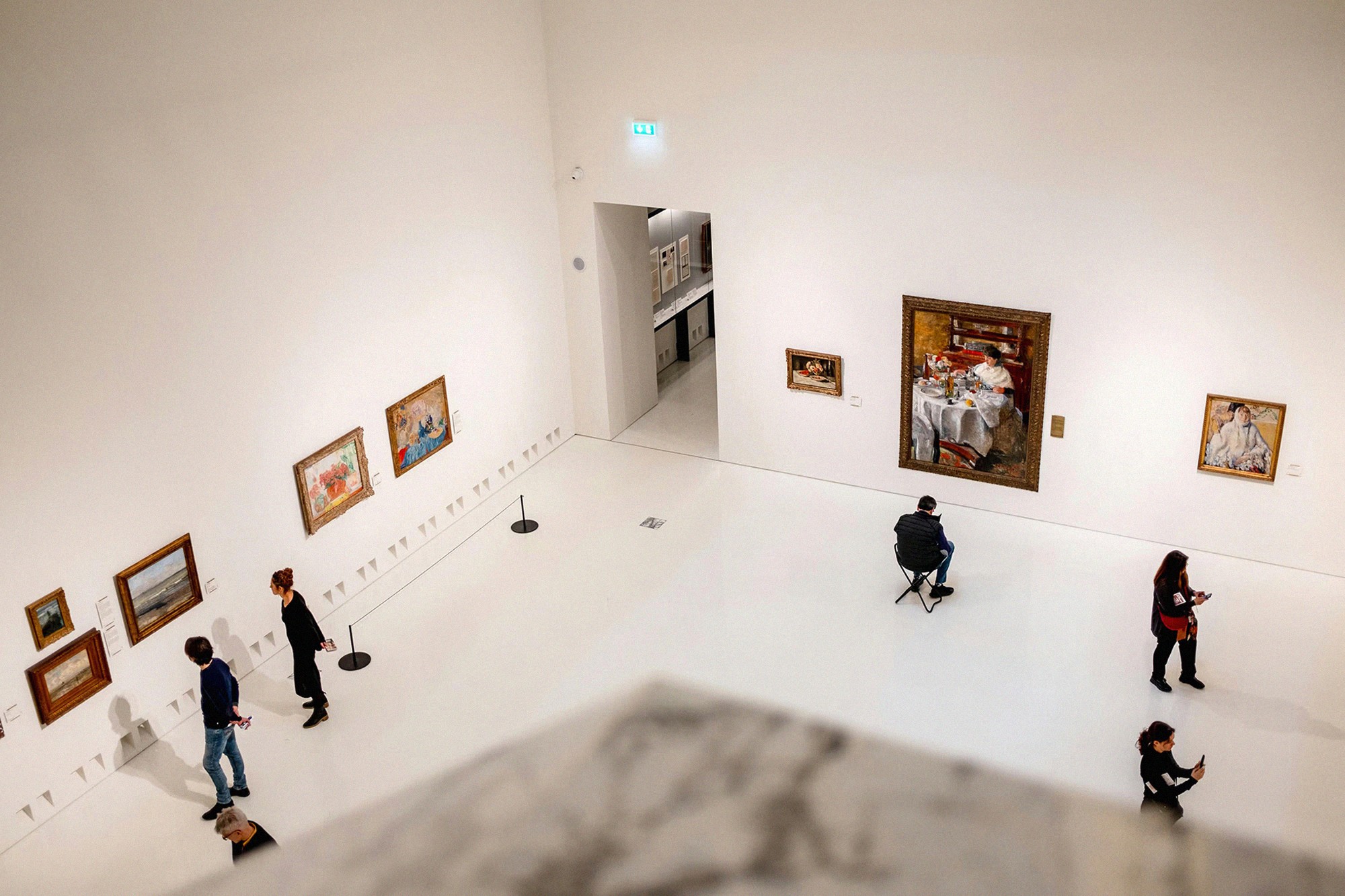 Wide overhead view of a contemporary art gallery with framed paintings and visitors exploring the room.
