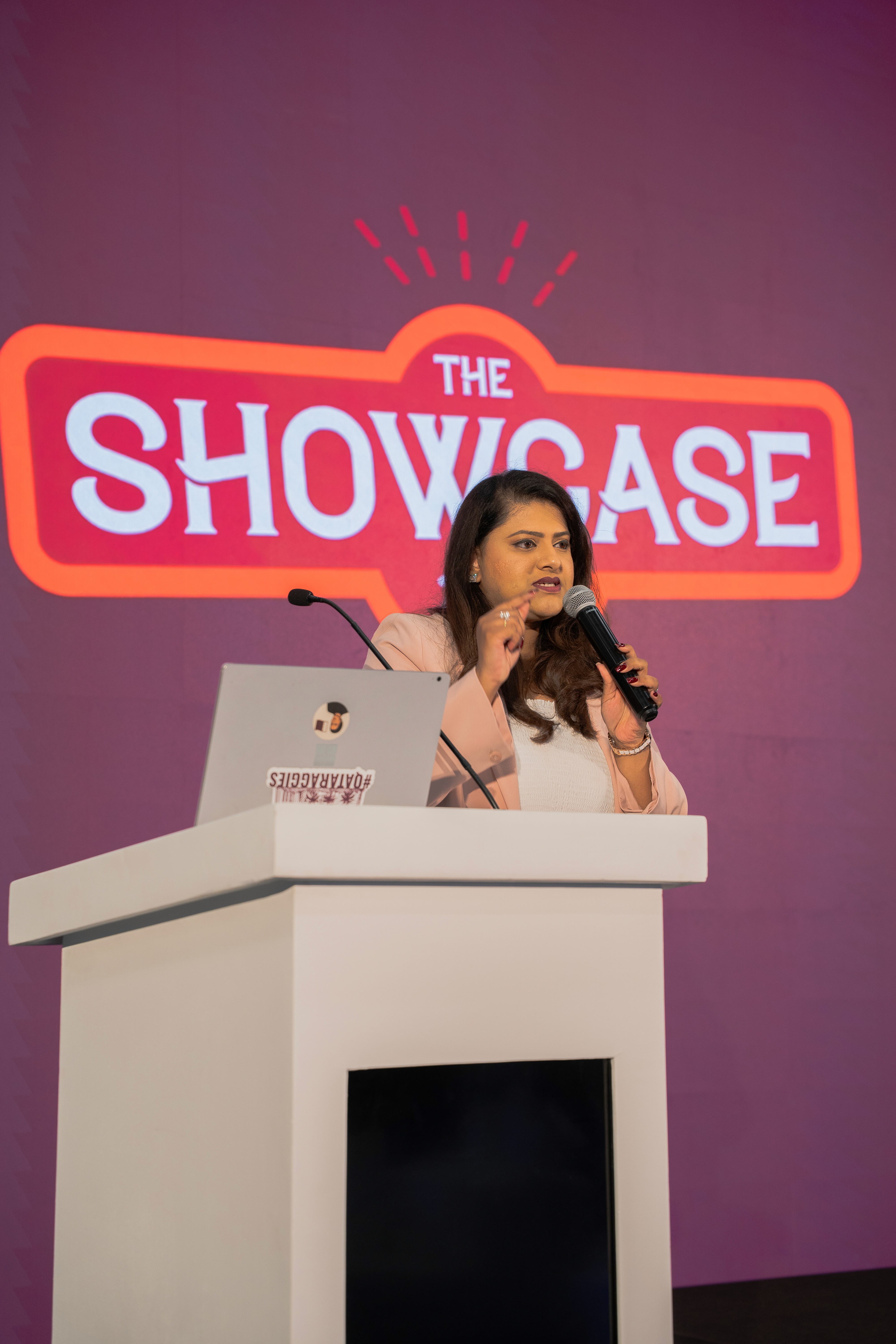 Smiling young woman with long hair standing against a dark green background, holding a finger to her chin.
