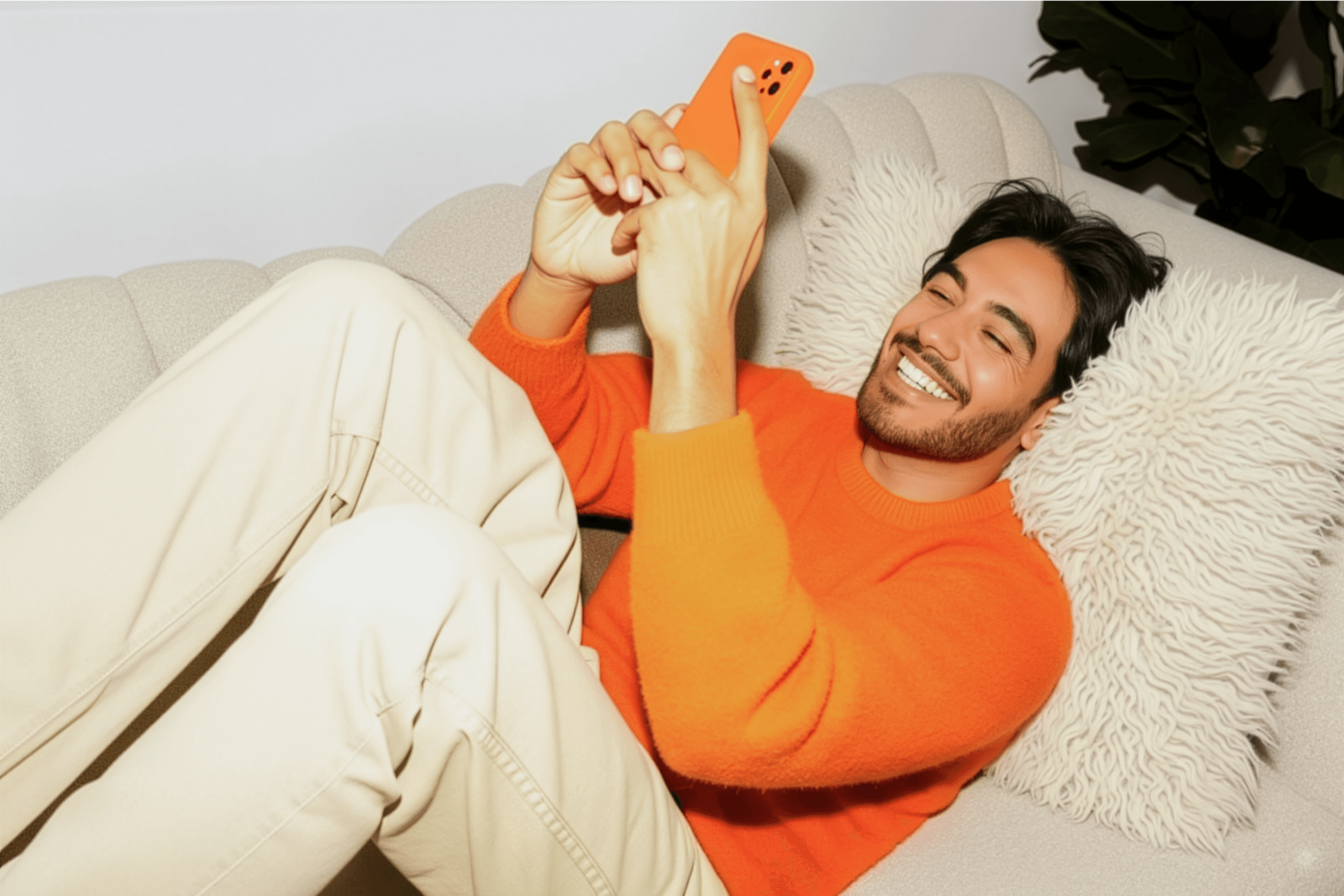 Hombre sonríe relajado en el sofá mientras mira emocionado su celular naranja.