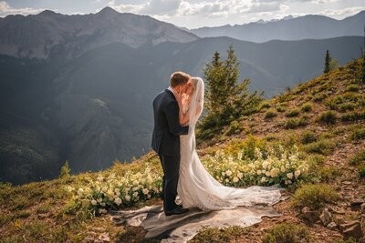 The couple kiss during their ceremony, surrounded by white flowers on the mountainside.