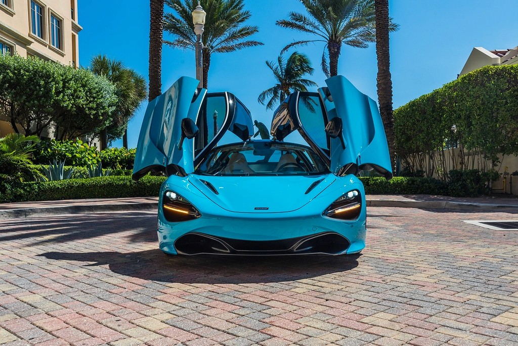 A stunning blue McLaren 720S with dihedral doors open, parked in a luxurious setting with palm trees and an elegant building in the background. This exotic supercar features a sleek design, powerful twin-turbo V8 engine, and unparalleled performance, making it the perfect choice for a luxury car rental experience.