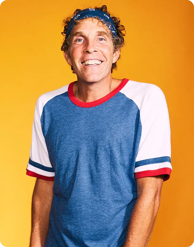 Smiling man with curly hair wearing a blue and white t-shirt and a headband, standing in front of a bright yellow background with a relaxed posture.