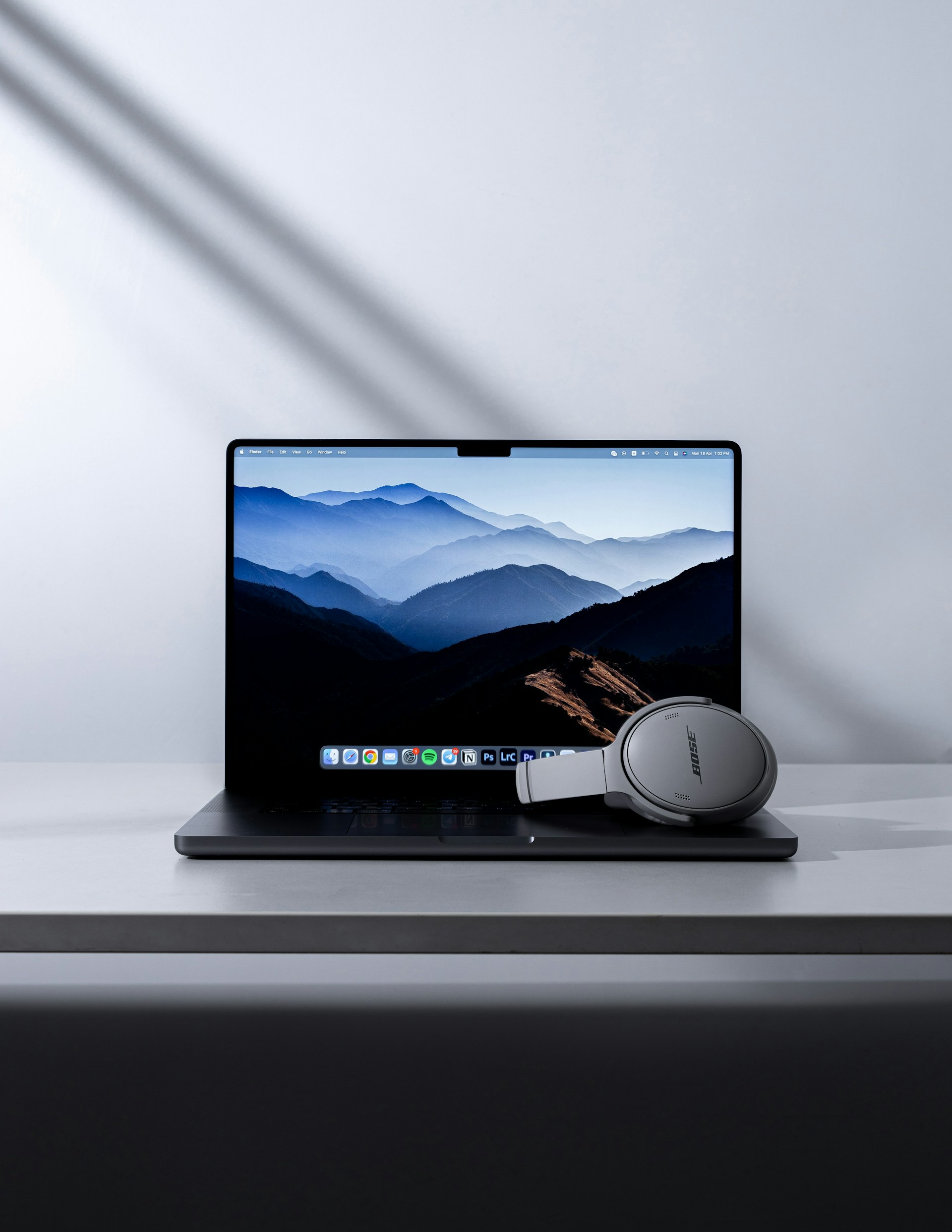 A laptop displaying a mountain landscape wallpaper is sitting on a desk with a pair of light gray over-ear headphones resting on the keyboard.