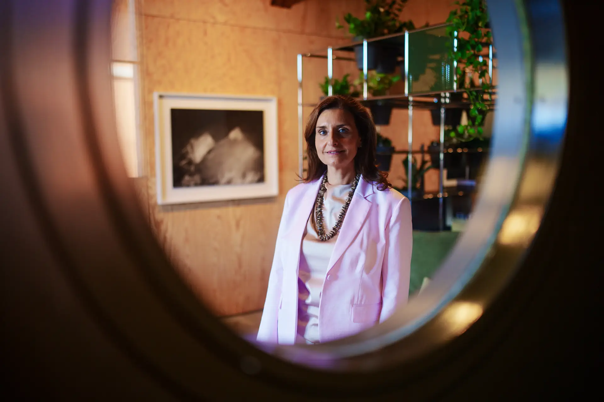 Porträt von Claudia Winiger im hellrosa Blazer, fotografiert durch einen runden Rahmen vor einem modernen Bürointerieur mit Pflanzen.