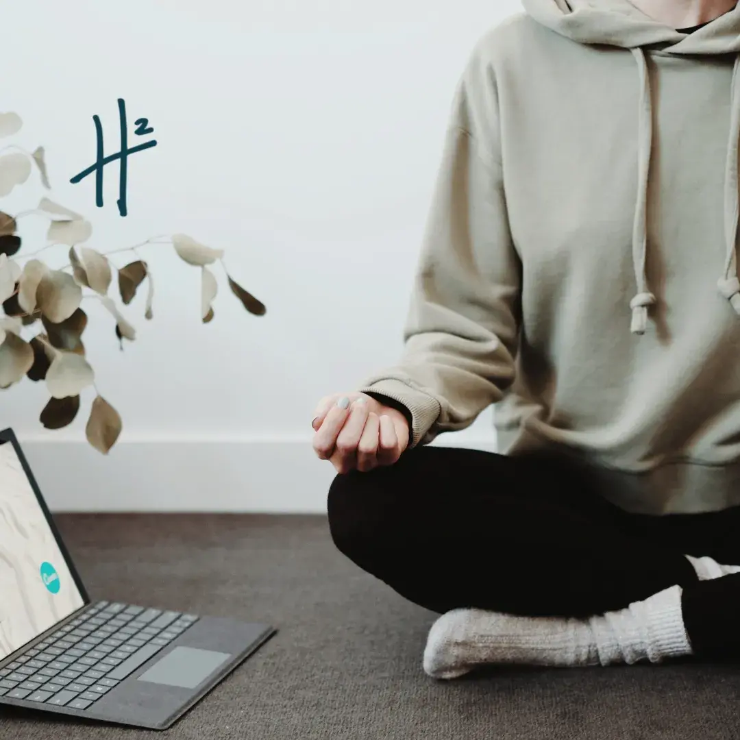 A person wearing a gray hoodie and black pants sits cross-legged on a carpeted floor, holding a cup, with a laptop nearby and a plant in the background, creating a cozy and relaxed atmosphere.