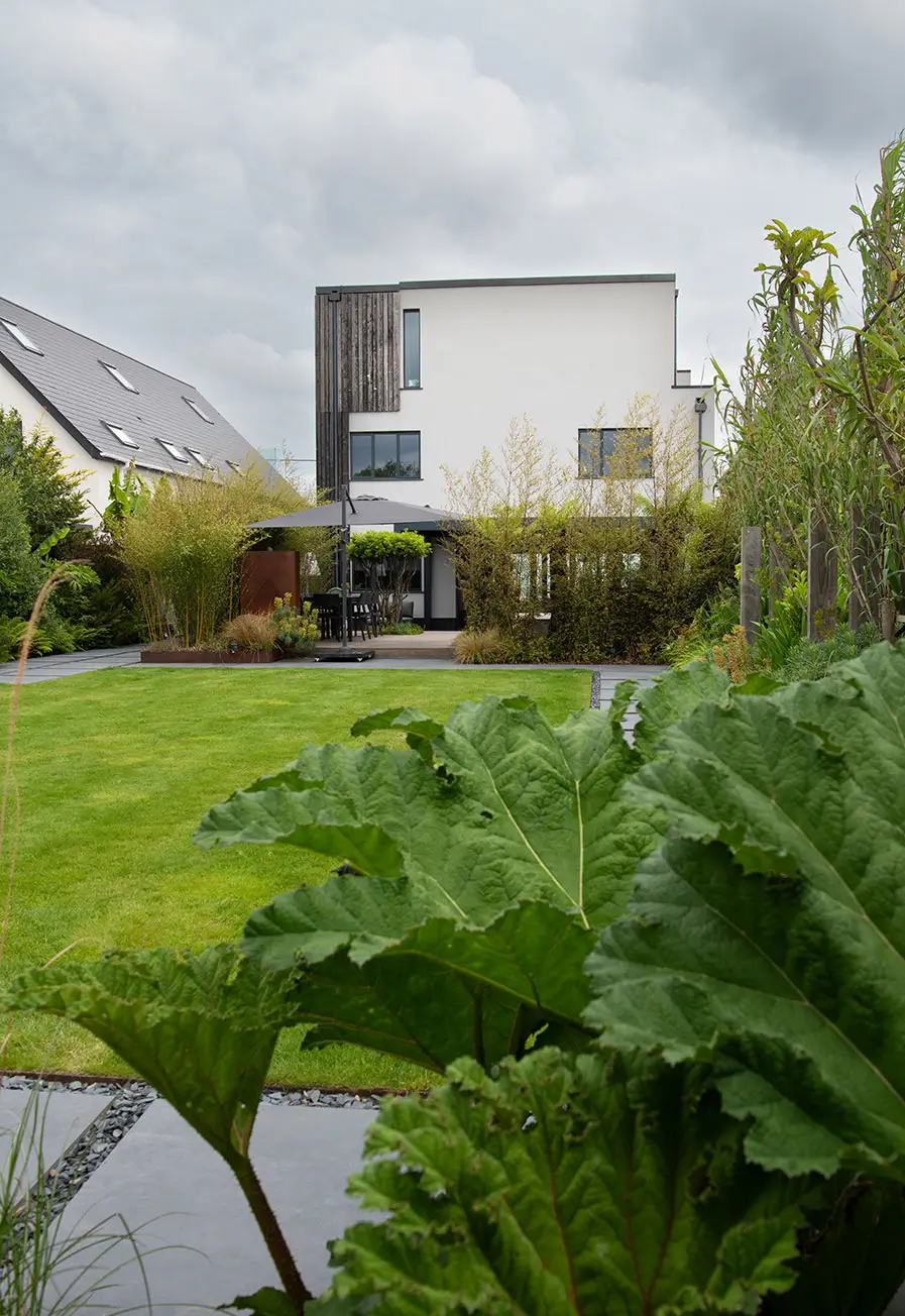A modern house with a sleek design sits behind lush green foliage under a cloudy sky.