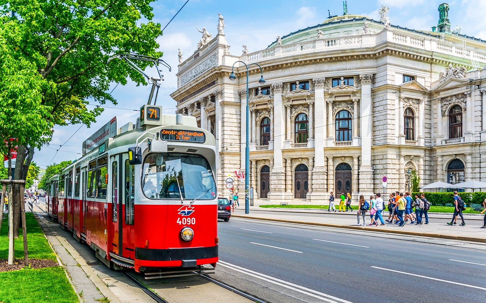 Red tram in front of Vienna's Burgtheater, Austria, with people walking nearby.