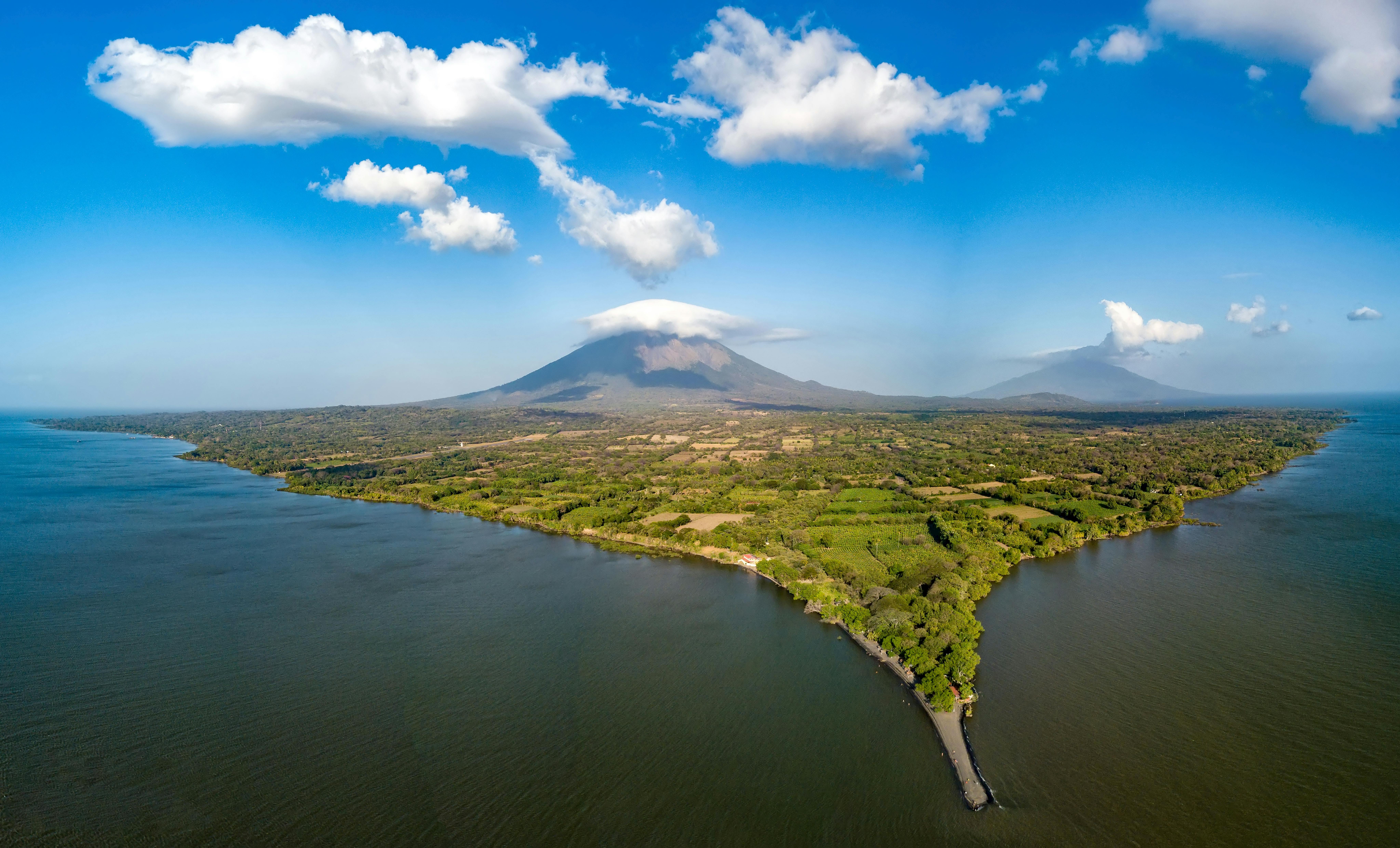An aerial view of Ometepe Island in Lake Nicaragua. The lush, green volcanic island is surrounded by dark water, with the towering Concepción Volcano visible in the center, its peak partially covered by a white lenticular cloud.