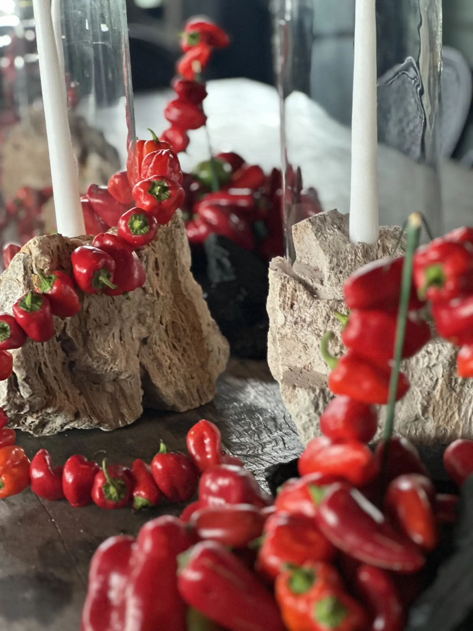 Detalle de instalación gastronómica con ajíes rojos pequeños, carbón vegetal y velas blancas en bases de piedra rugosa.