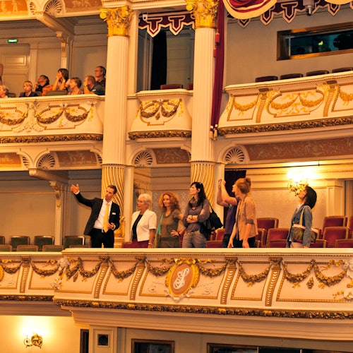 Ihmiset seisovat ja istuvat koristeellisilla teatteriparvekkeilla, jotka on koristeltu kultaisilla yksityiskohdilla ja seppeleillä, tarkkaillen salia.