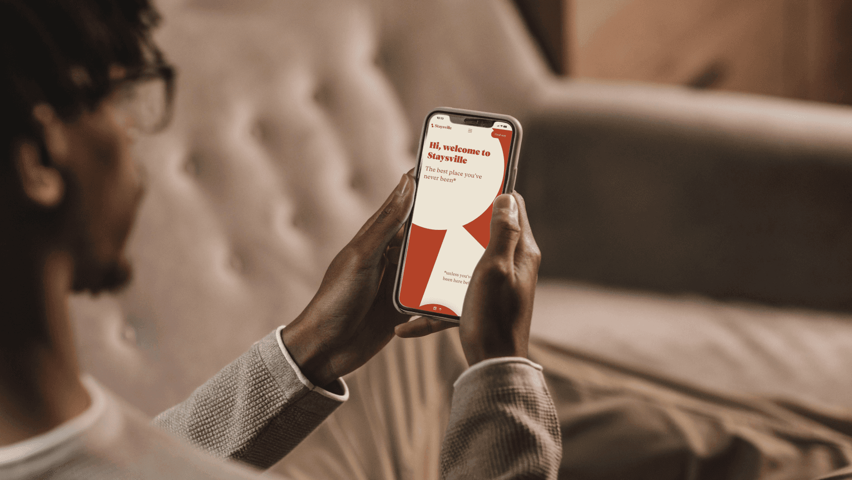 A man sitting on a brown sofa holding an iPhone showing the frontpage of the Staysville webpage.