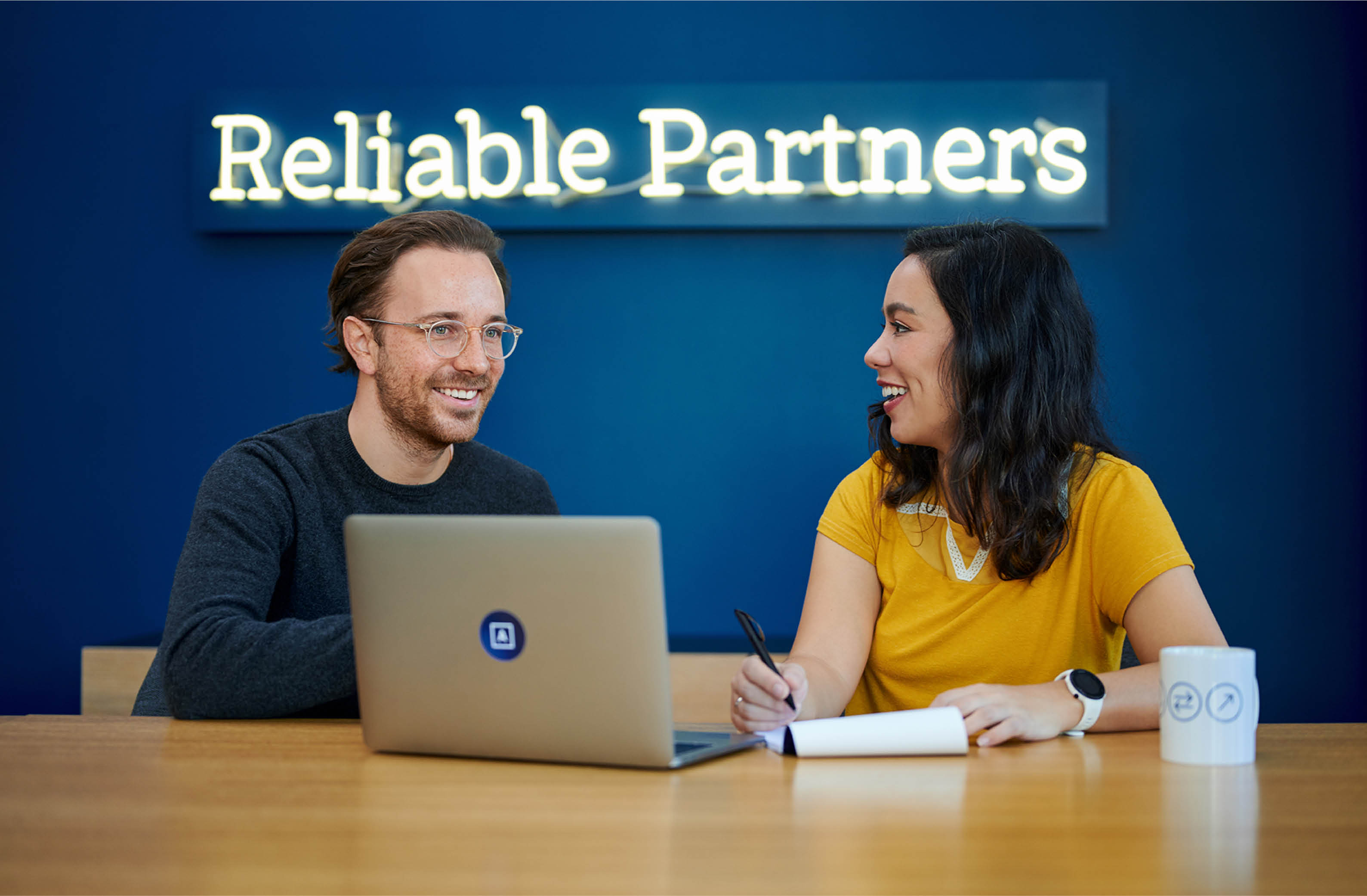 A man and woman engaged in a collaborative discussion at a table with a laptop, under a sign that says 'Reliable Partners,' ideal for showcasing teamwork in a professional environment.