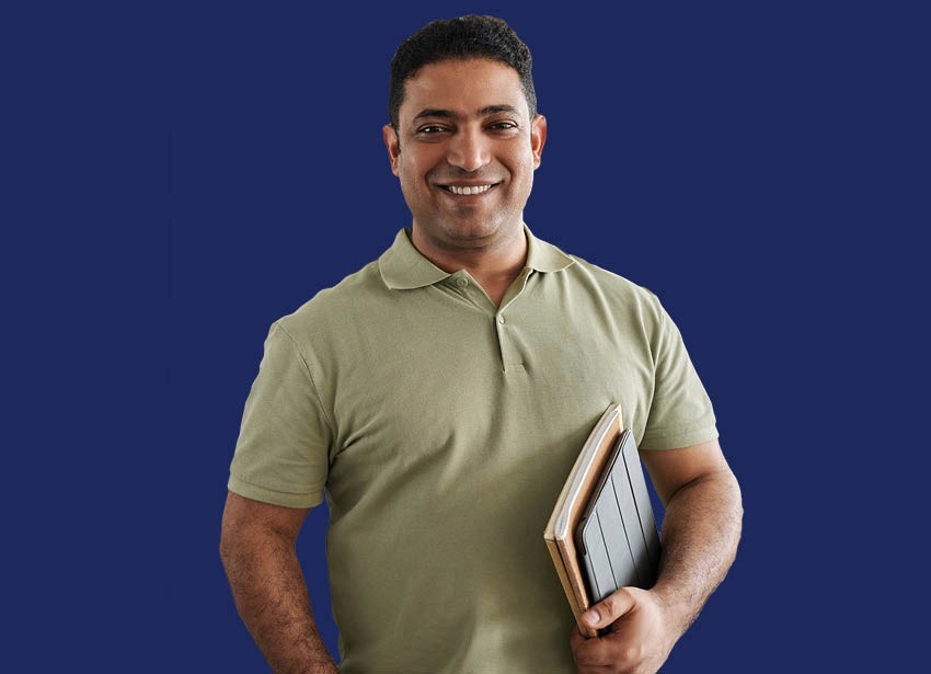 A man in a sage green shirt holding a tablet and books shown in front of a navy background