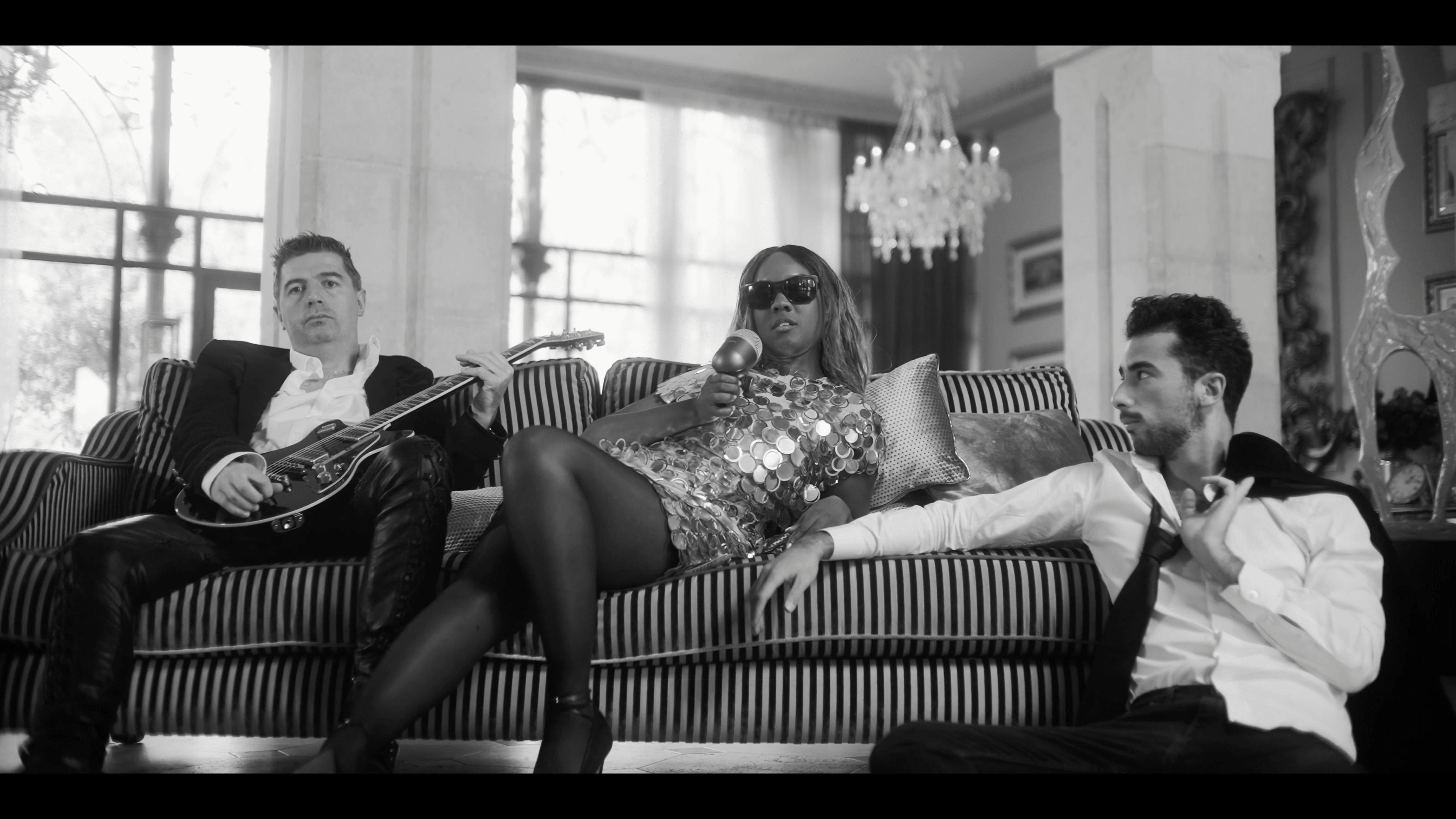 Three people are seated on a striped sofa in an elegant room, with two men and one woman in a shimmering dress, as one man holds a guitar; a chandelier and large windows are visible in the background.