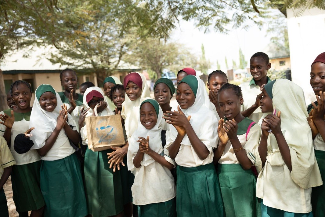 A group of smiling schoolgirls in green and white uniforms stand outdoors under leafy trees, joyfully clapping and holding a handmade project, embodying education and community spirit in an open-air setting.