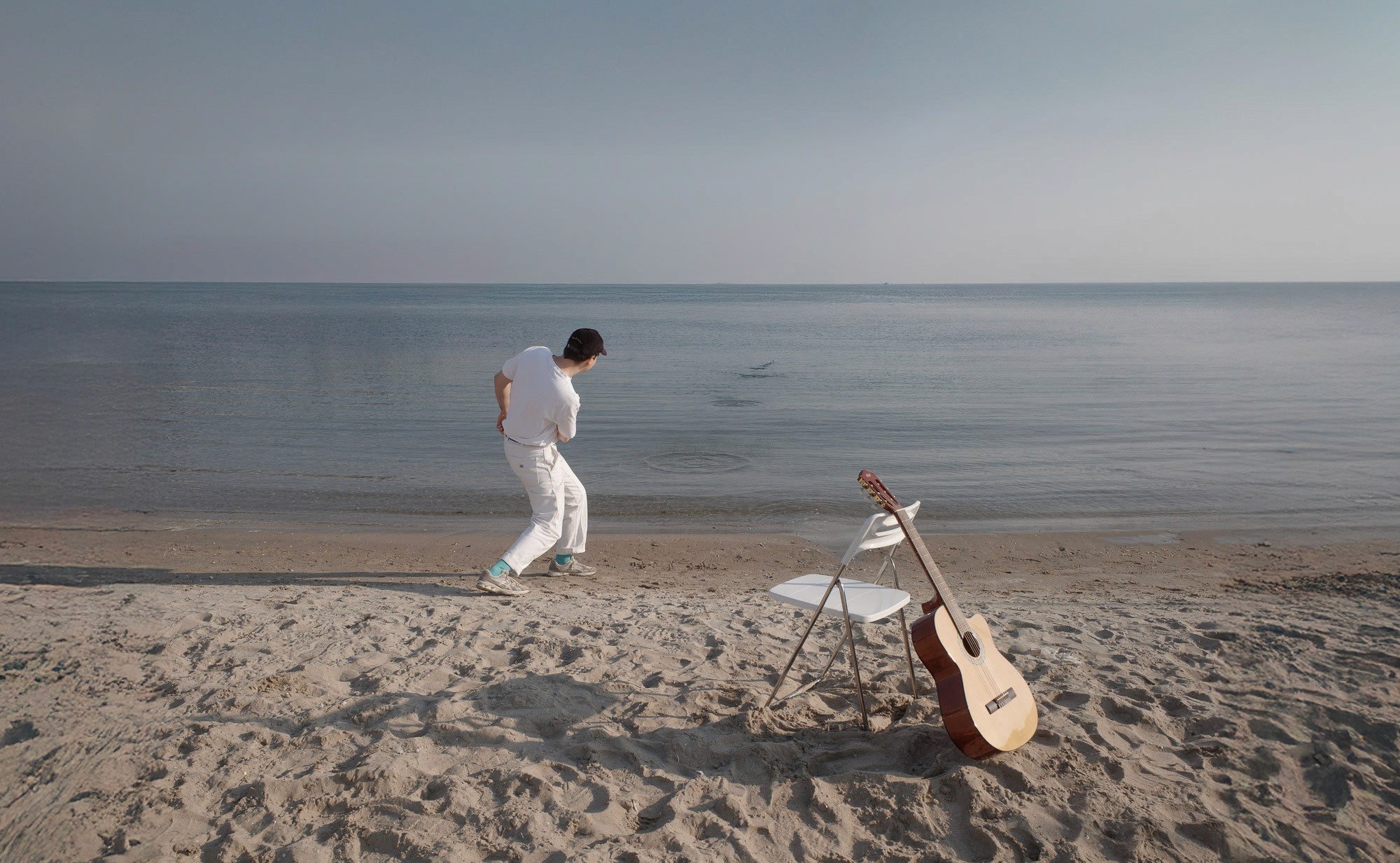 A person dressed in white dances on a serene, sandy beach with a calm sea in the background, while a guitar leans against a white chair nearby, capturing a peaceful coastal vibe.