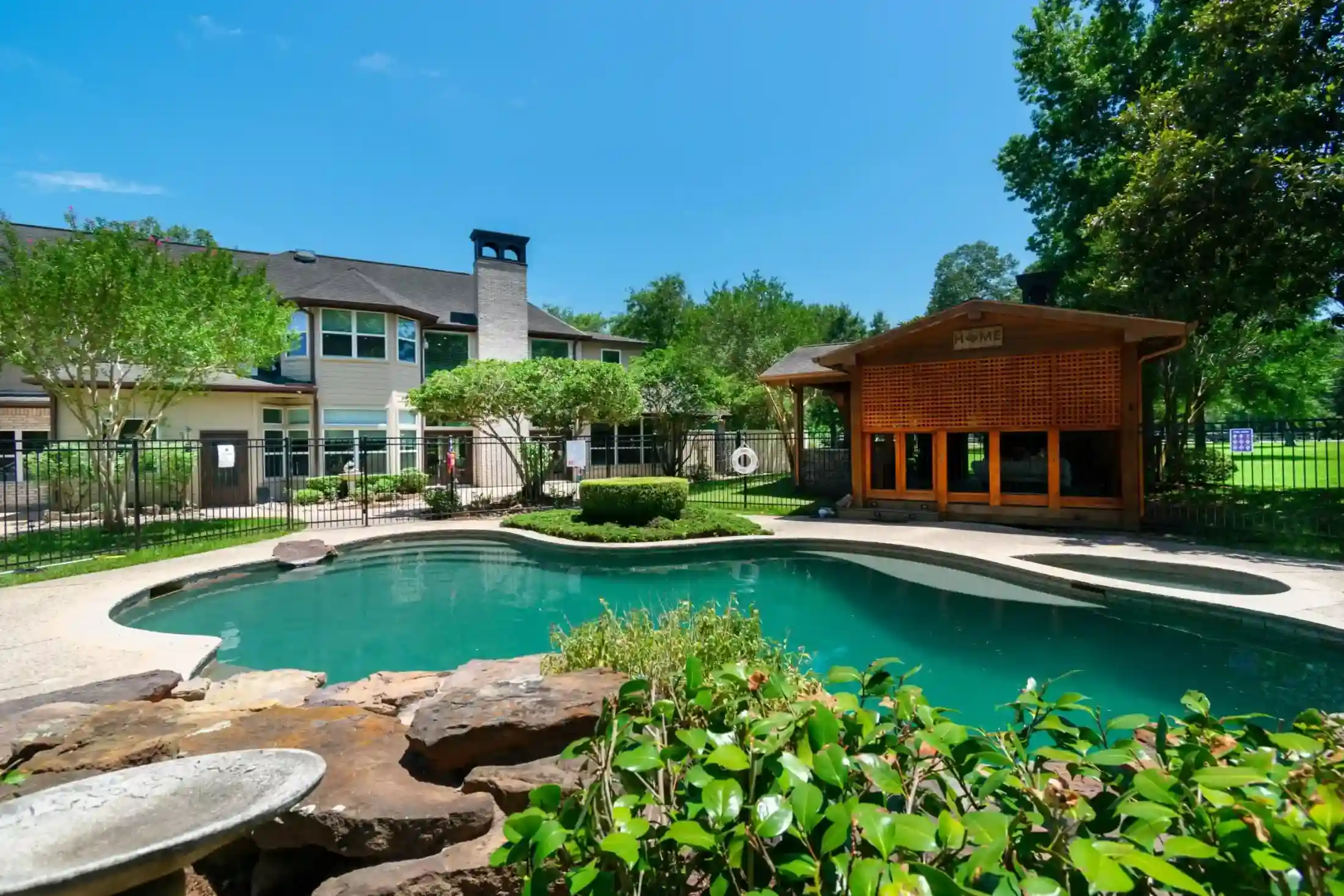 The backyard of the Monte Nido Facility with a curved swimming pool, a stone border, green plants, a wooden gazebo, and a two-story house in the background, surrounded by trees and a black iron fence under a clear blue sky.