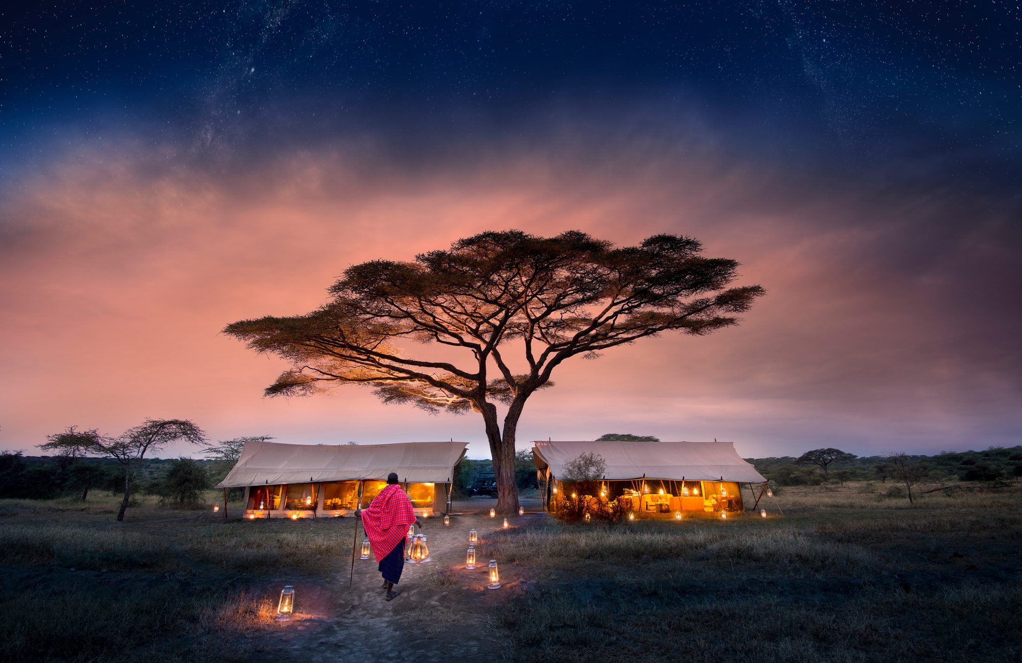 A Maasai man in a red shuka walks a lantern-lit path toward two glowing canvas tents beneath an acacia tree at dusk in Tanzania's Serengeti.