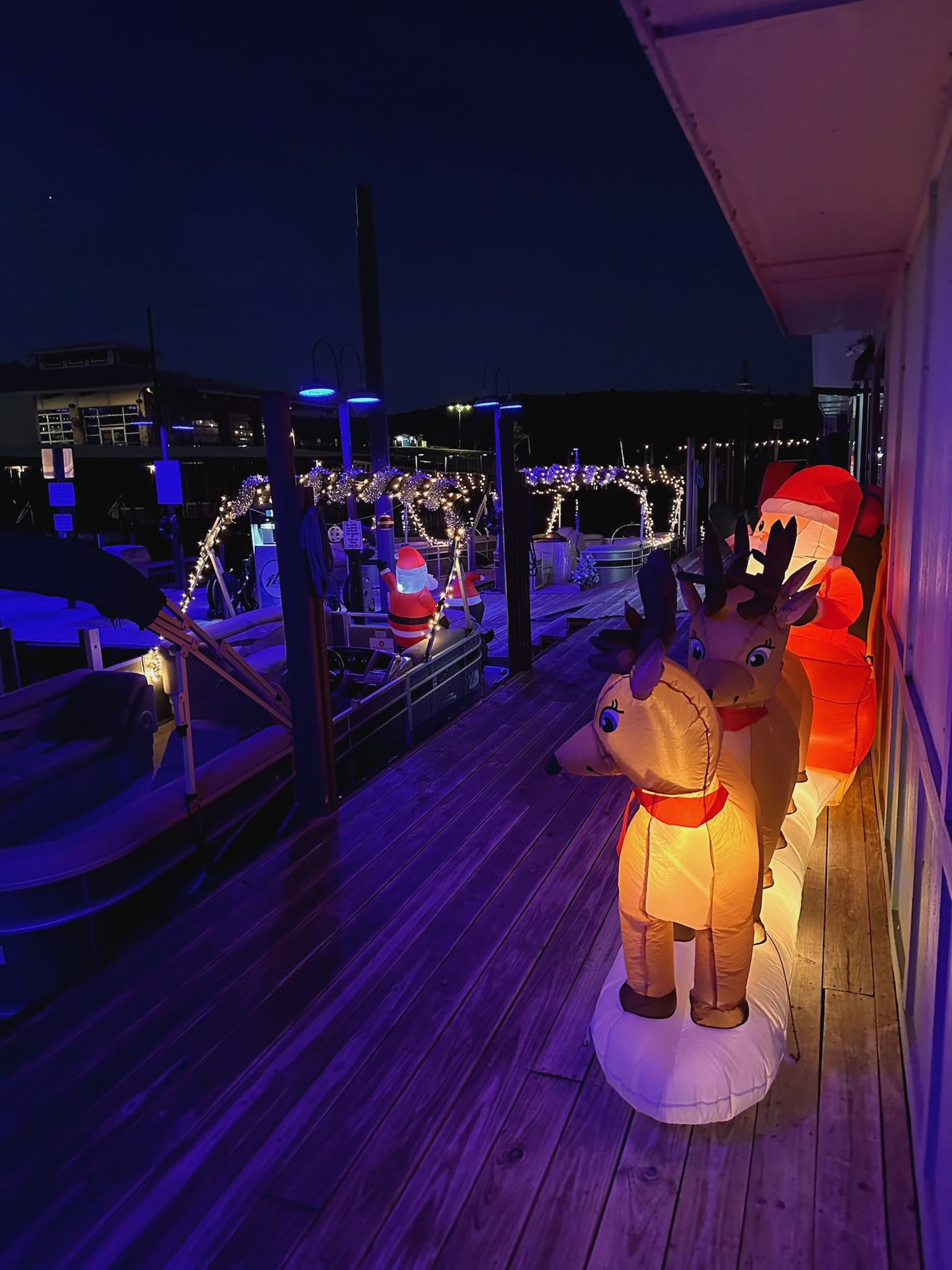A festive dock scene at night featuring a light-up inflatable display of Santa and reindeer, surrounded by boats decorated with string lights, creating a warm and cheerful holiday atmosphere.