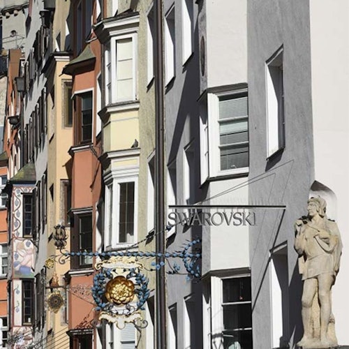 Rua com edifícios coloridos e estreitos, uma placa de loja da Swarovski, placas suspensas ornamentadas e uma estátua de um homem à direita.