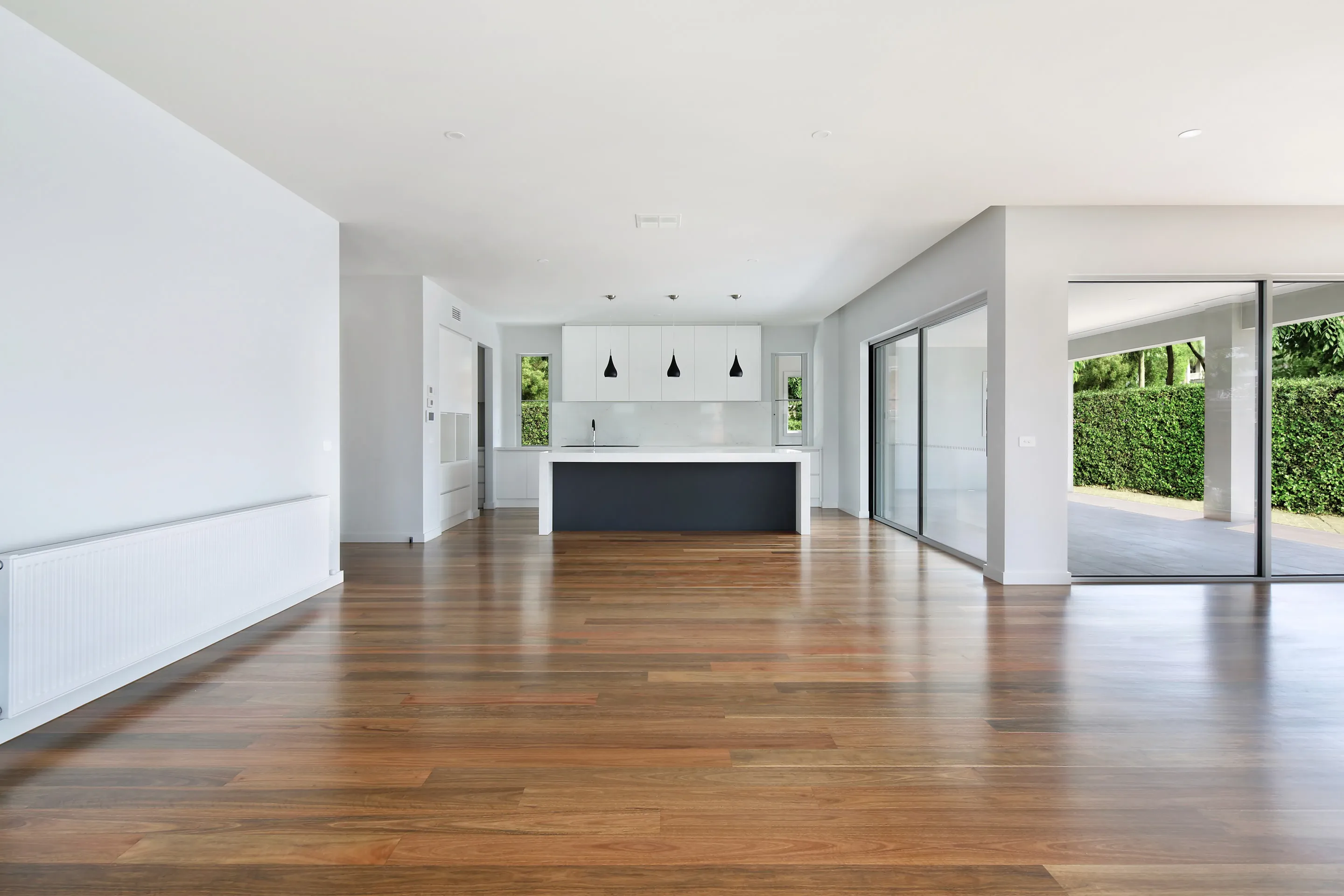 Open-plan living and kitchen with polished timber floors, a white kitchen with navy island, black pendant lights and sliding doors to a covered terrace and hedge