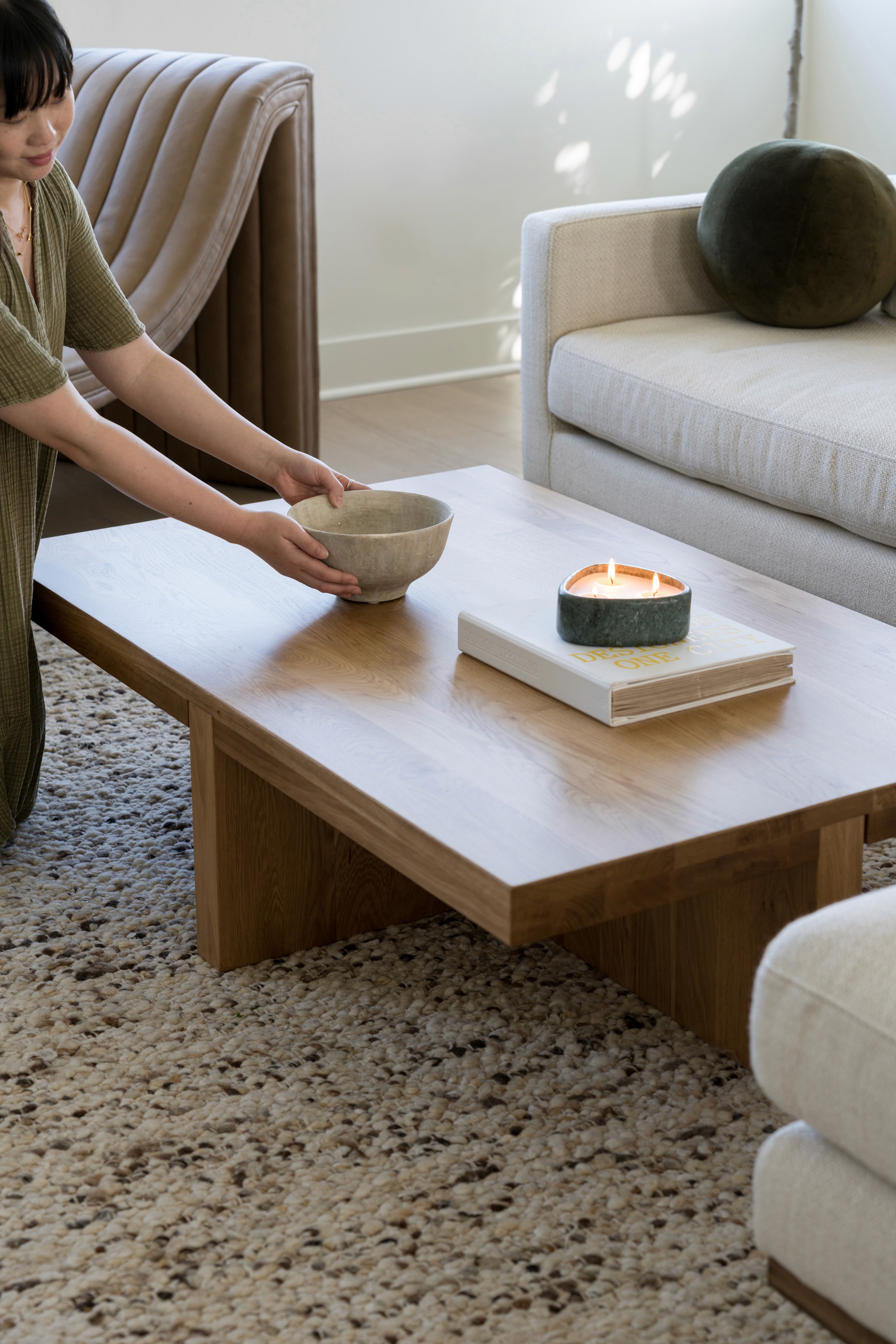 Light wood coffee table with ceramic bowl, large coffee table book, and green marble candle against woven rug