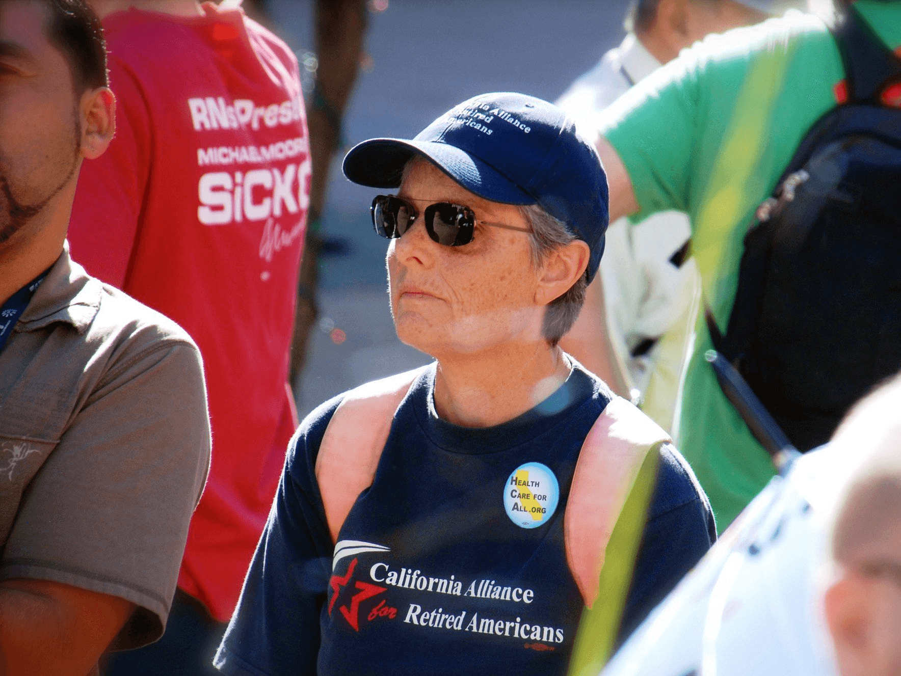 A person wearing sunglasses, a cap, and a “California Alliance for Retired Americans” shirt stands attentively among a crowd at a public event or rally.