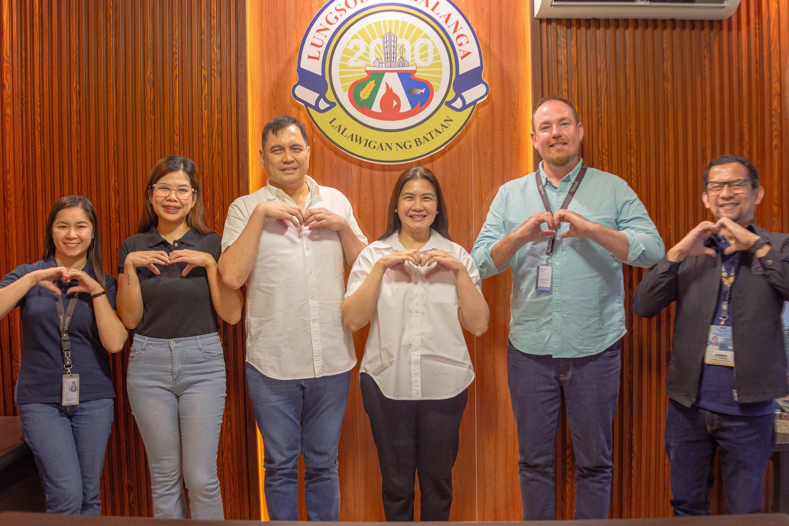 A diverse group of seven people stand smiling in front of a wooden wall with a seal, posing with hand gestures.