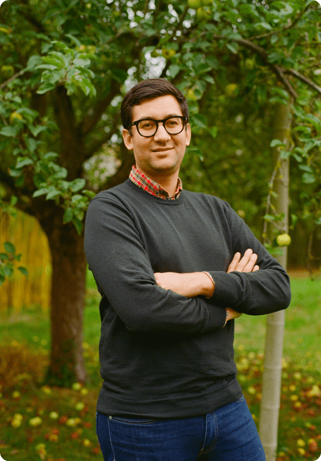Portrait d'un homme souriant portant des lunettes et un pull noir, posant les bras croisés devant un pommier dans un jardin.