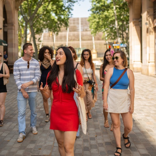 Grupa ljudi u šetnji na otvorenom tijekom sunčanog dana, s predvodnicom u crvenoj haljini koja razgovara s okupljenima. U pozadini se vide drveće i zgrade.