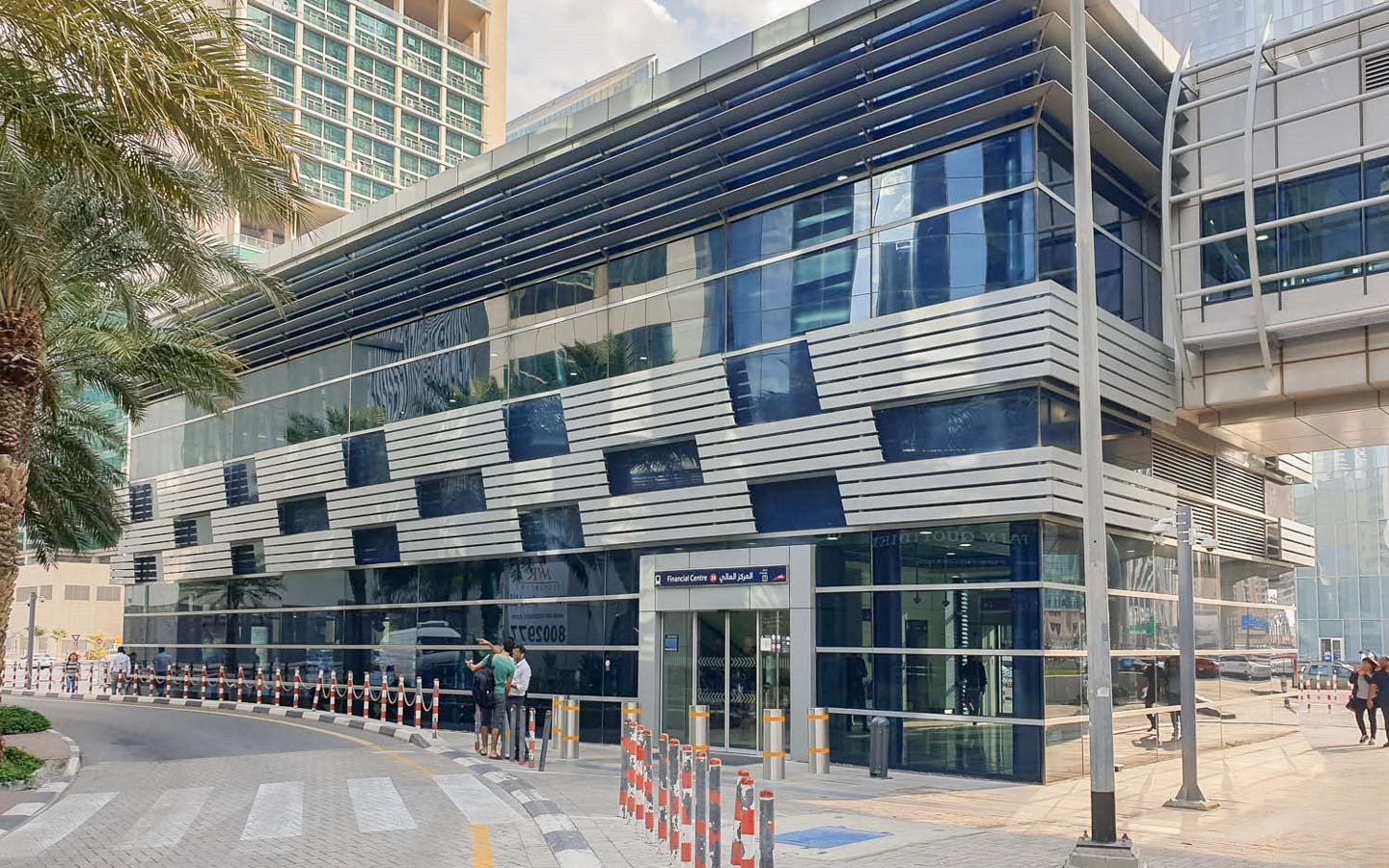 The glass and silver-toned façade of the modern Dubai Design District metro/tram station building with palm trees nearby.
