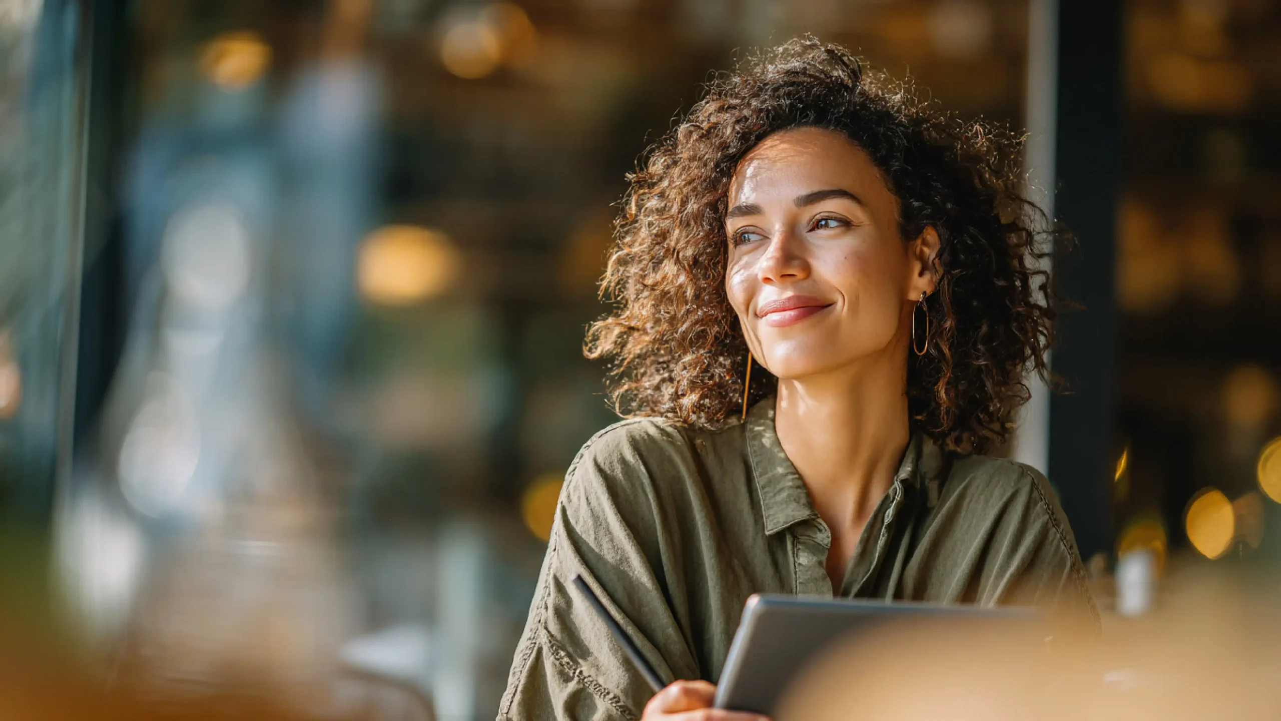 A cheerful woman with curly hair smiles while sitting at a table in a cozy, warmly lit café.
