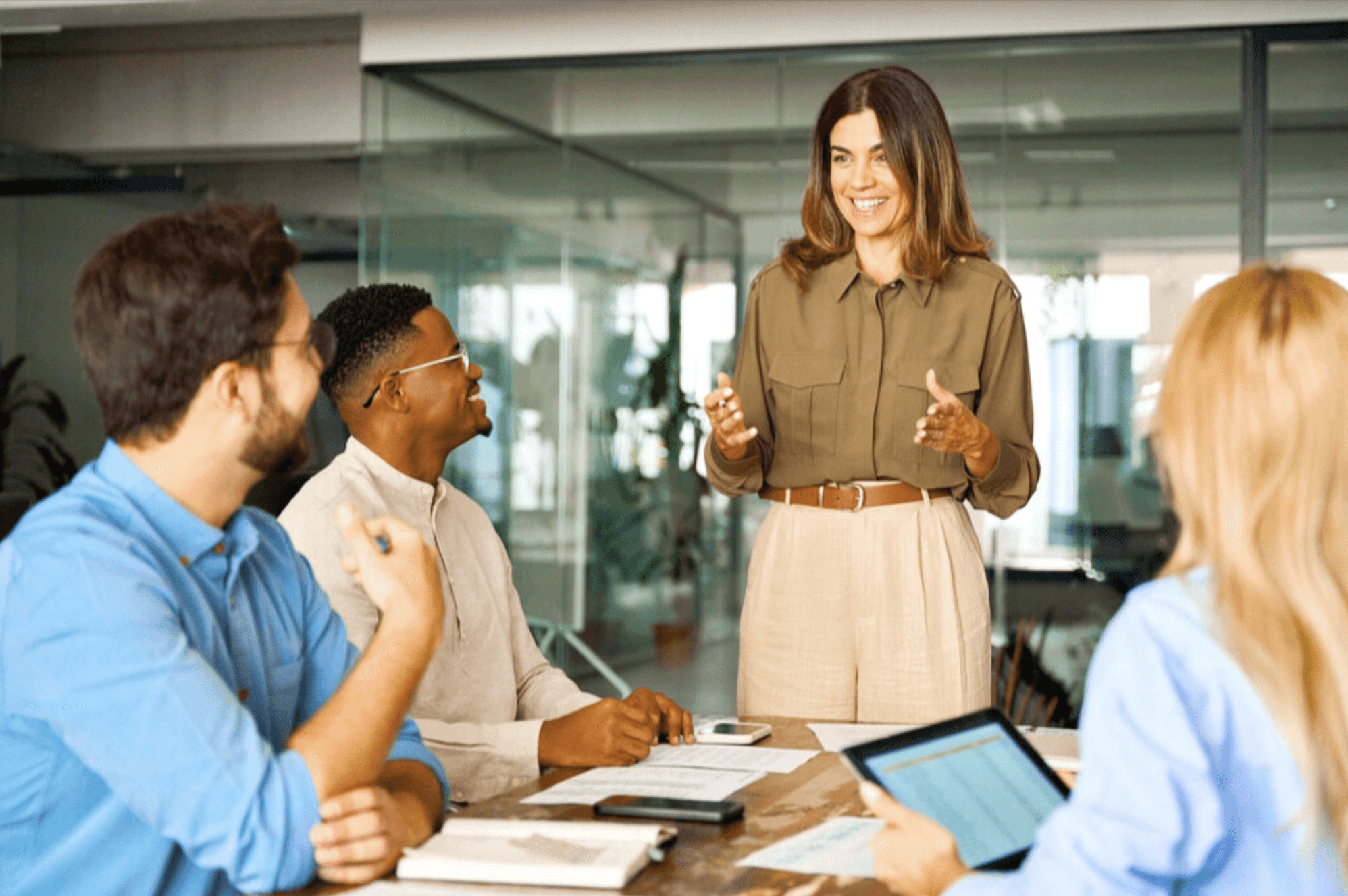 A woman in an olive green shirt stands and leads a meeting, gesturing with her hands while three colleagues sit and listen at a wooden table in a bright, modern office.
