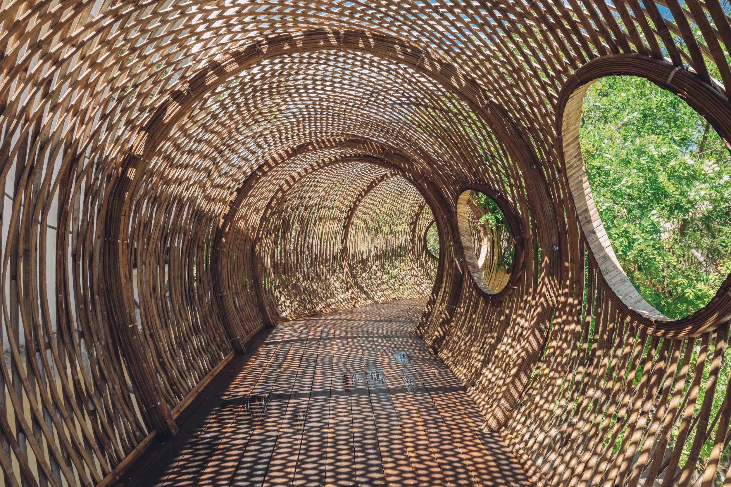 Interior perspective of the Cervecería Tulum bamboo tunnel, where the woven lattice skin creates intricate light and shadow patterns on the wooden walkway.