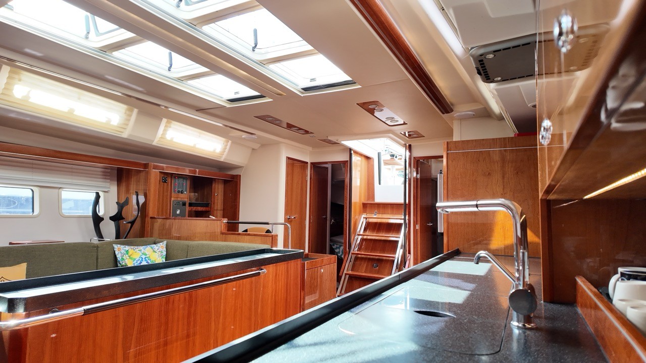 Interior view of the Hanse 575's saloon and galley, showing rich mahogany cabinetry, a granite countertop with a chrome faucet, an olive-green settee, overhead skylights, and companionway stairs leading below.