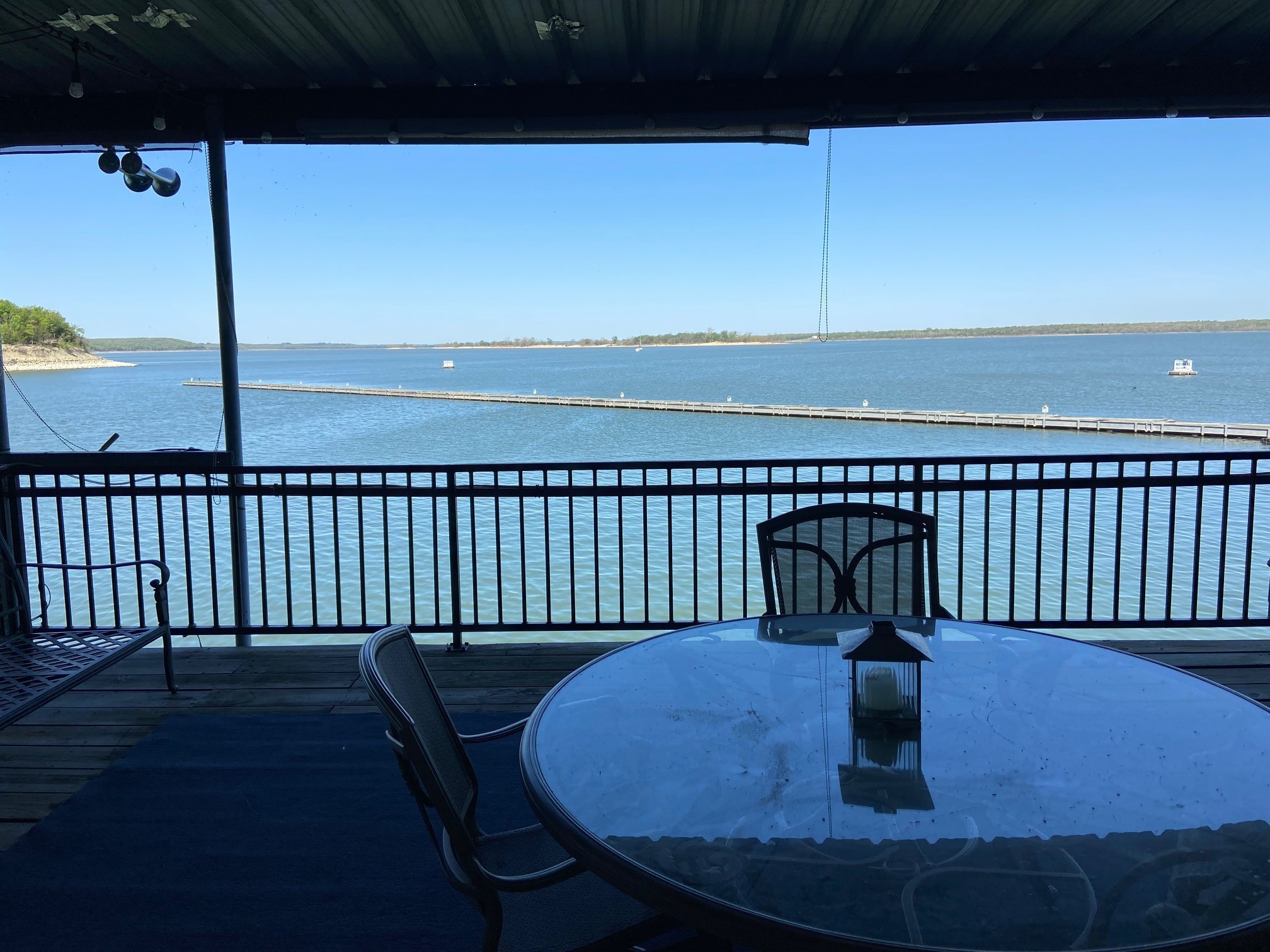 A tranquil lake view from a covered deck features a glass table and chairs on a wooden floor, framed by a railing, with clear blue skies and calm waters stretching into the horizon, providing a perfect setting for outdoor relaxation.