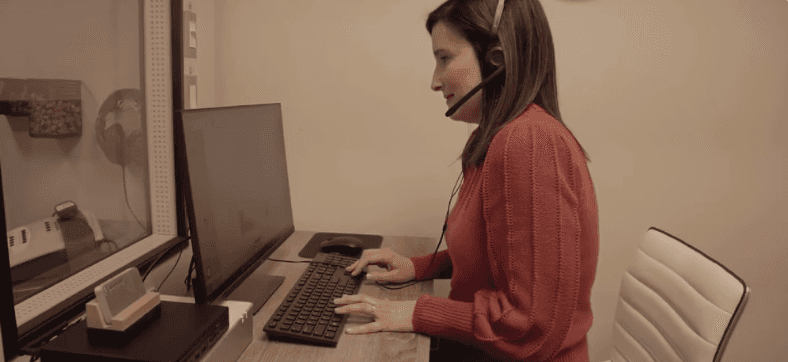 A woman with short gray hair sits on a couch, holding her cheek, appearing to be in discomfort.