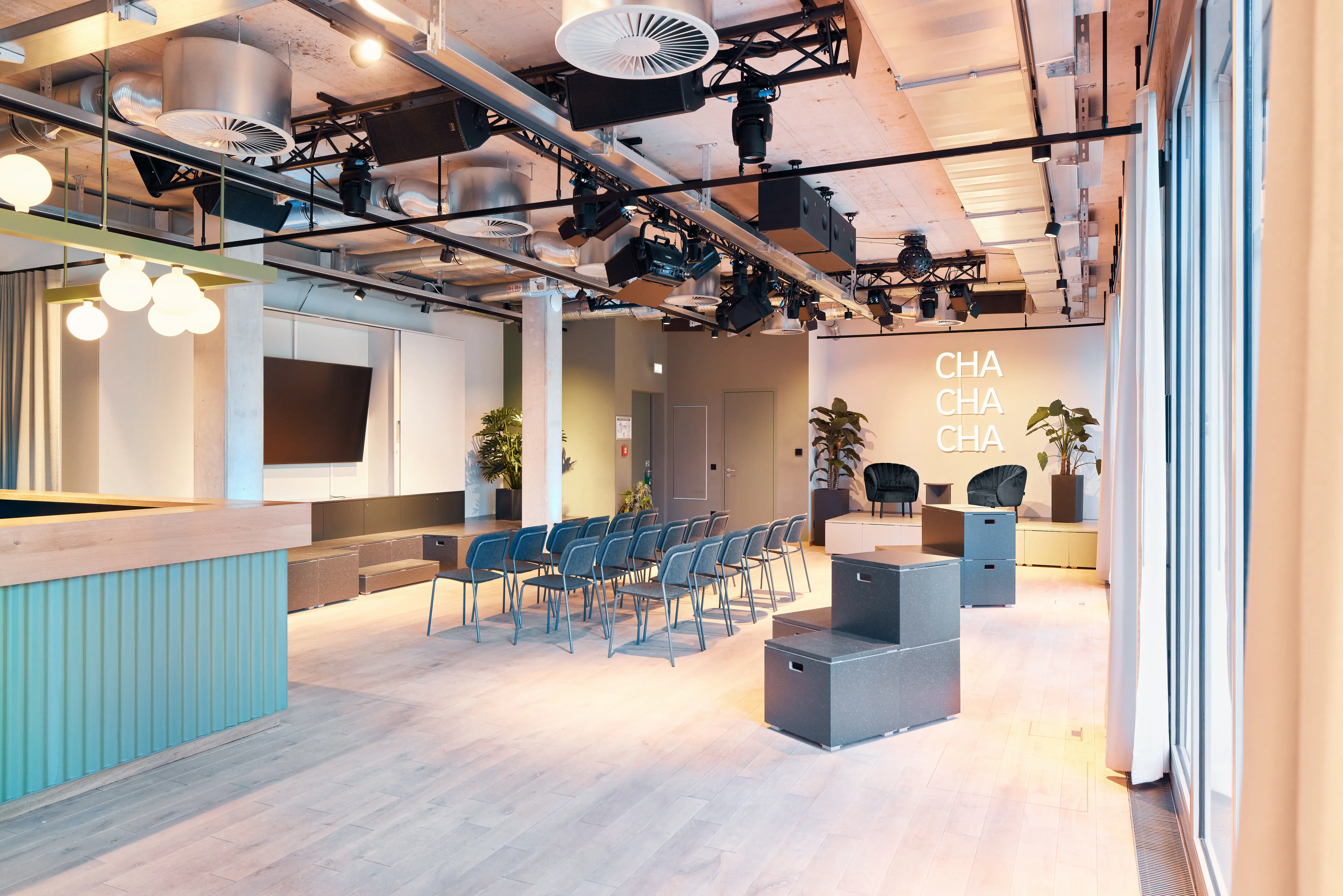 Event space at 381 EVENT SPACE with turquoise corrugated bar counter, wall-mounted screen, blue chair rows, grey seating boxes and stage lighting on black trusses.