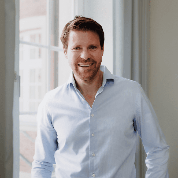 Jüngerer männlicher Geschäftsführer im hellen Hemd steht entspannt vor einem Fenster im Büro von SLB.