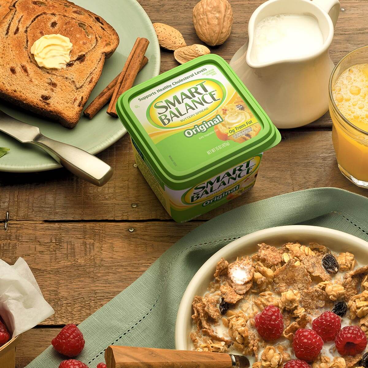 Breakfast scene with Smart Balance spread, a glass of orange juice, a bowl of cereal with raspberries, buttered toast, a jug of milk, and nuts on a wooden table.
