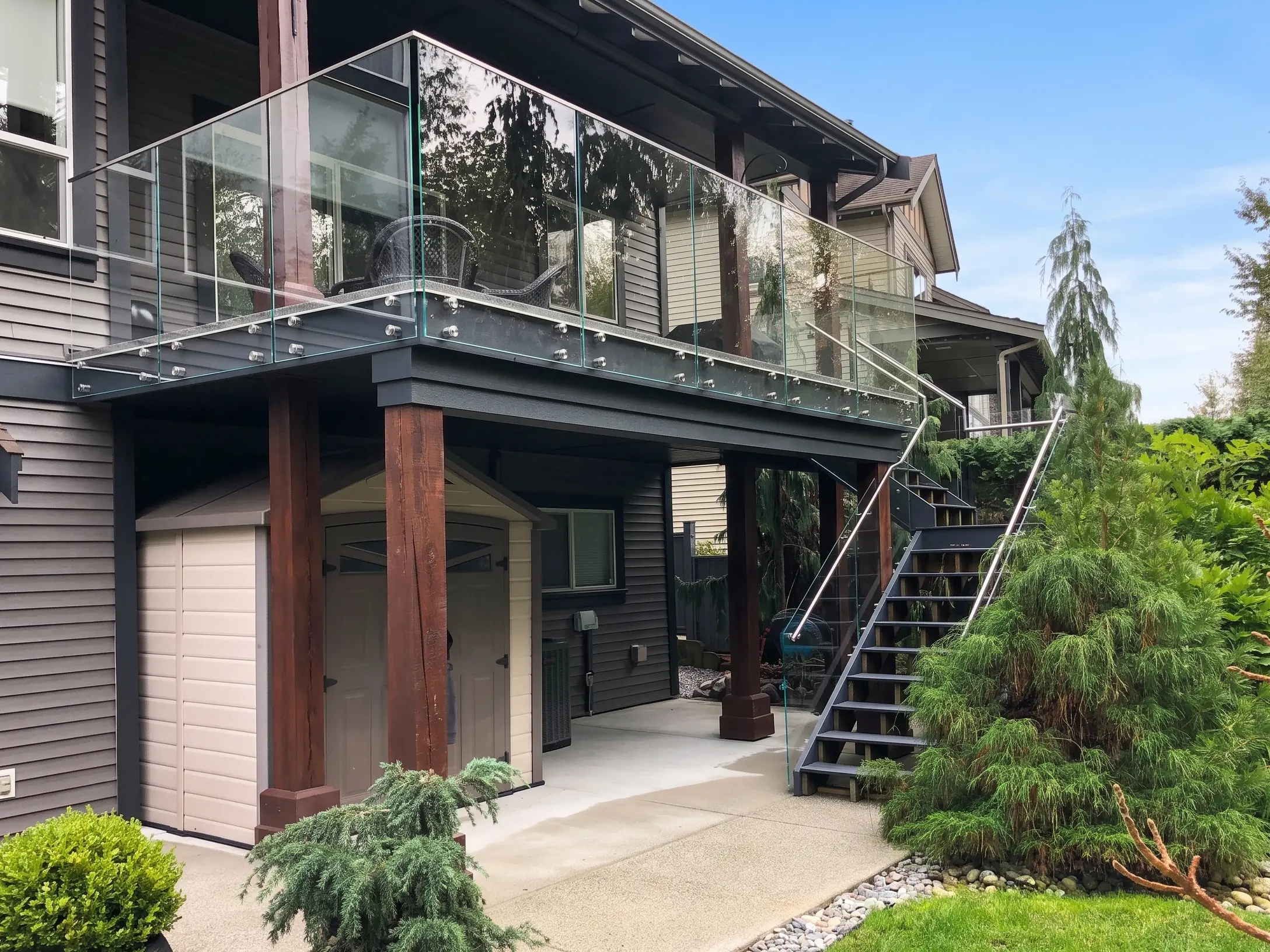 Two-storey Vancouver Island home with frameless glass railings on the upper deck and a glass-railed exterior staircase, set against landscaped gardens.
