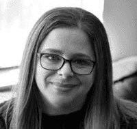 Michelle, in a black and white photo, a white woman with long brown hair wearing glasses and black top smiling at the camera