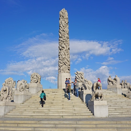 People climb stairs surrounded by stone sculptures, leading to a tall monolith against a blue sky with scattered clouds.